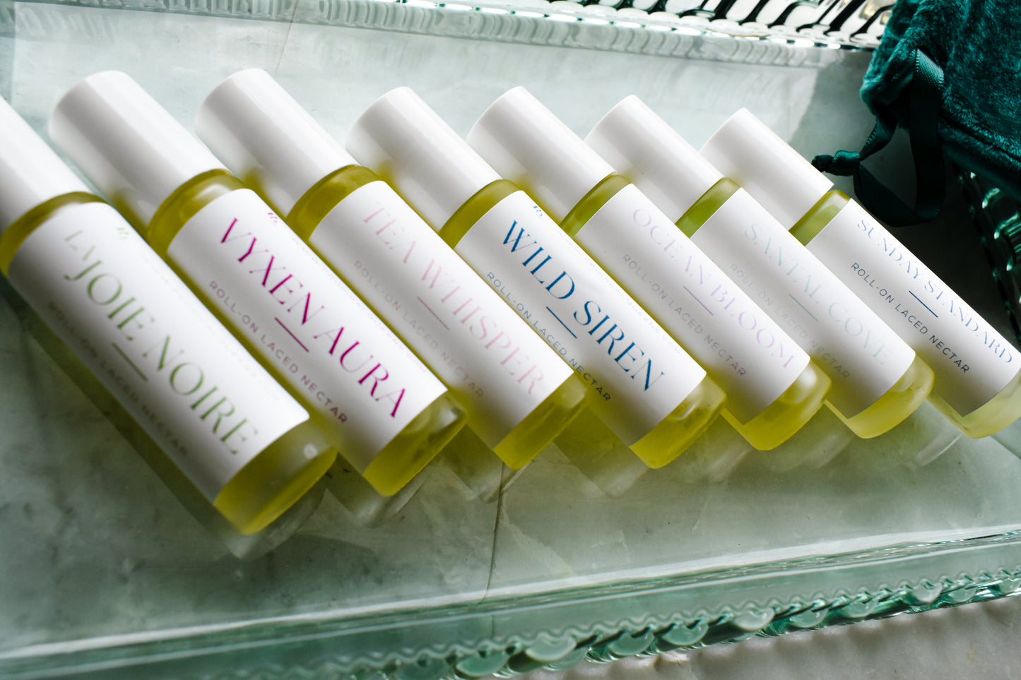 Six small cylindrical cosmetic bottles filled with pale yellow liquid are arranged diagonally on a clear glass tray. Each bottle has a white cap and a white label with different colored text, i