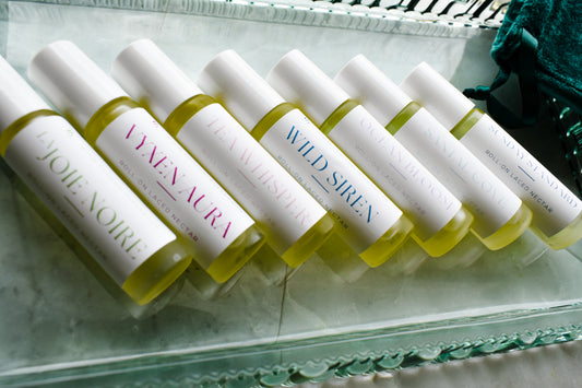 Six small cylindrical cosmetic bottles filled with pale yellow liquid are arranged diagonally on a clear glass tray. Each bottle has a white cap and a white label with different colored text, i