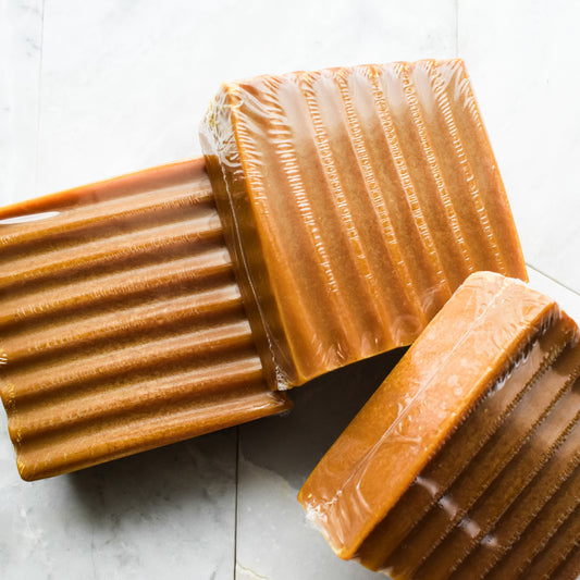 Three rectangular bars of caramel-colored soap wrapped in clear plastic, arranged on a light gray marble surface.