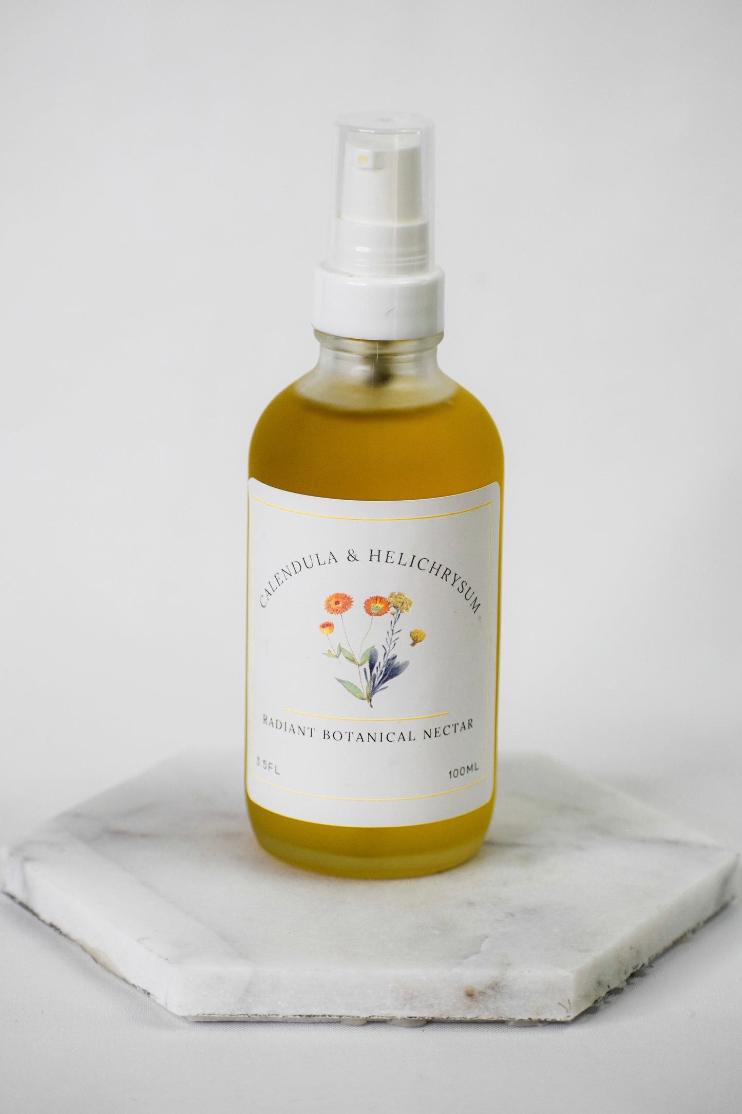 A glass spray bottle filled with golden yellow liquid, featuring a white label with botanical illustrations of flowers and the text 'Pleiodula & Helichrysum Radiant Botanical Nectar'. The bottle is placed on a white marble hexagonal coaster against a clean, light gray background.