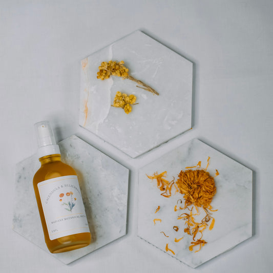 A bottle of botanical essential oil spray with a white label and clear cap, placed on a hexagonal marble tray next to dried yellow flowers on two other hexagonal marble trays against a light gray background.