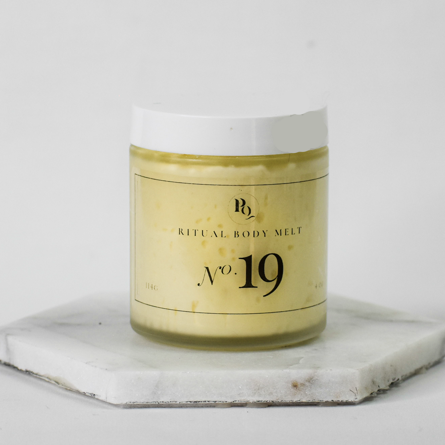 A clear glass jar of Ritual Body Melt No. 19 body butter with pale yellow wax inside and a white lid, displayed on a white marble hexagonal coaster against a light gray background.
