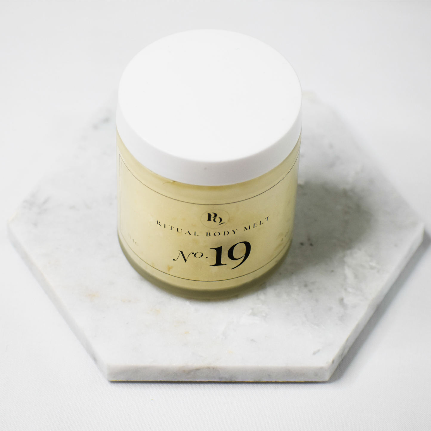 A jar of Ritual Body Melt No. 19 body melt, featuring a white lid and a pale yellow-beige body, displayed on a hexagonal white marble coaster against a clean white background.