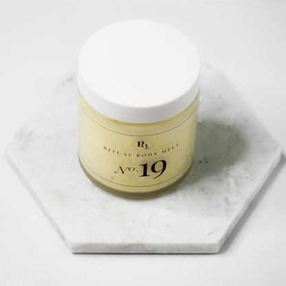 A jar of Ritual Body Melt No. 19 body melt, featuring a white lid and a pale yellow-beige body, displayed on a hexagonal white marble coaster against a clean white background.