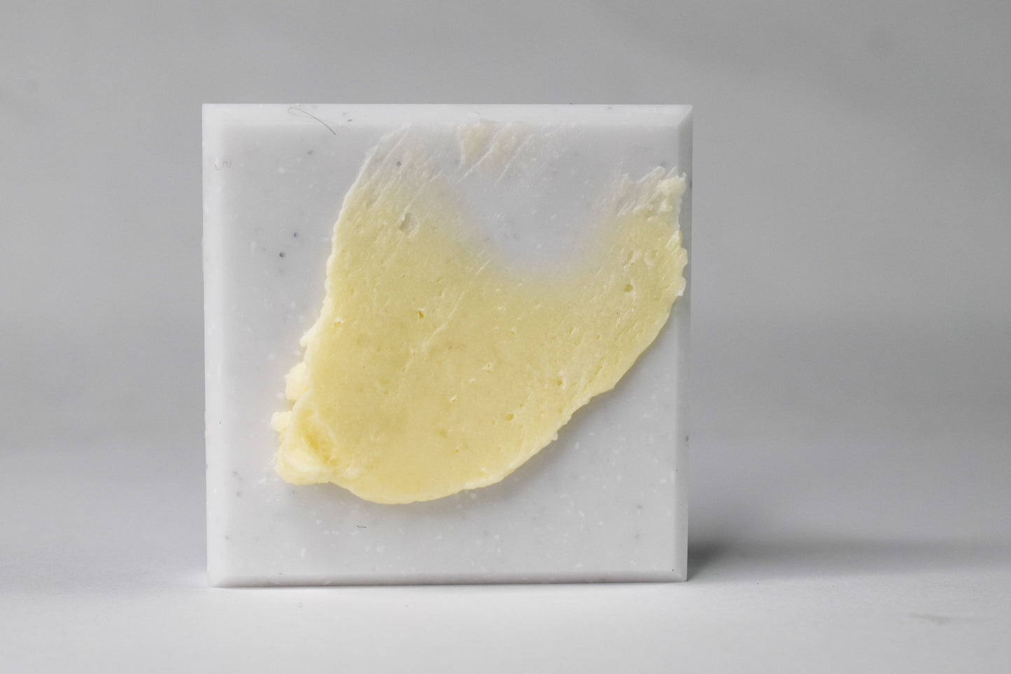 A square white marble soap bar with a textured, swirled pale yellow cream or butter-like substance embedded in the center, set against a neutral gray background.