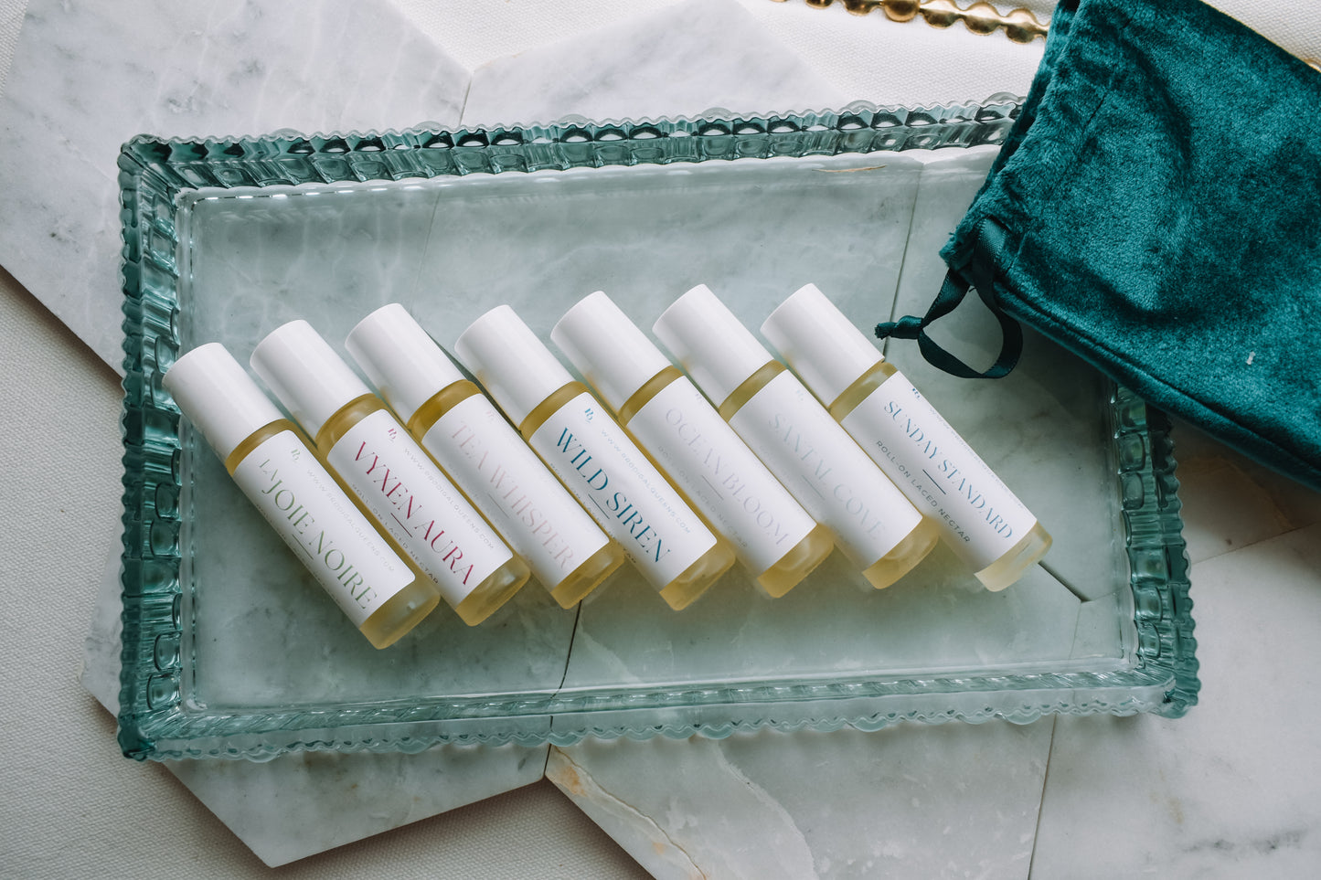 Six small cylindrical roller bottles with white labels and pale yellow liquid, arranged on a clear glass tray with a teal velvet drawstring bag beside them on a light marble surface.