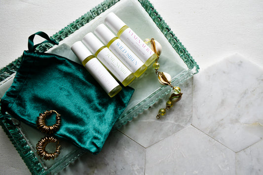 Four white roll-on bottles with yellow-green liquid and blue text labels, arranged on a clear glass tray with a teal satin pouch and gold jewelry on a light marble surface.