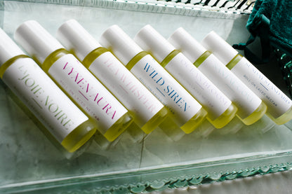 Six small cylindrical cosmetic bottles filled with pale yellow liquid are arranged diagonally on a clear glass tray. Each bottle has a white cap and a white label with different colored text, i