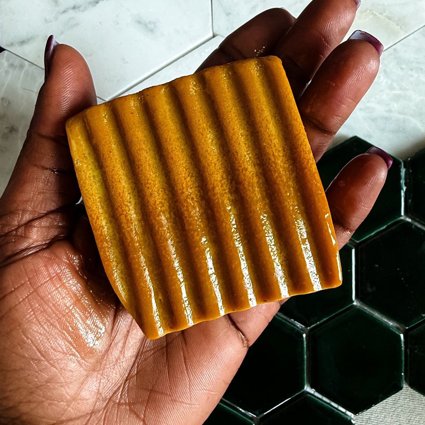 A hand holding a rectangular bar of golden-yellow soap with a ribbed texture, set against a background of white marble and dark green hexagonal tiles.