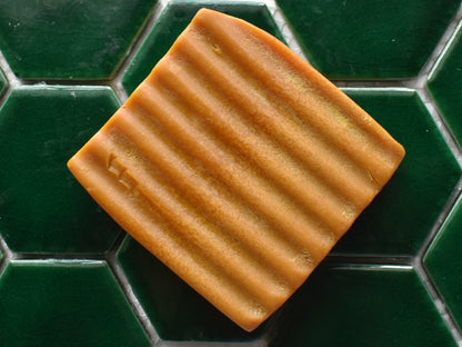 A rectangular bar of textured, ribbed soap in a warm caramel brown color, resting on a dark green hexagonal tiled surface.