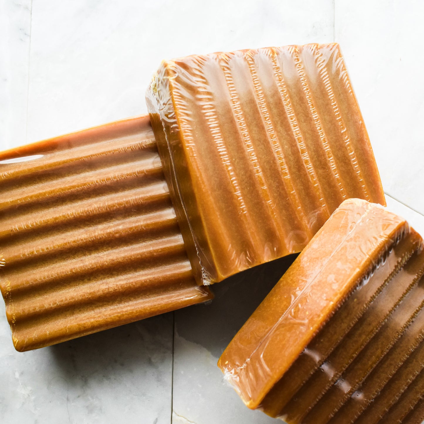 Three rectangular bars of caramel-colored soap wrapped in clear plastic, arranged on a light gray marble surface.
