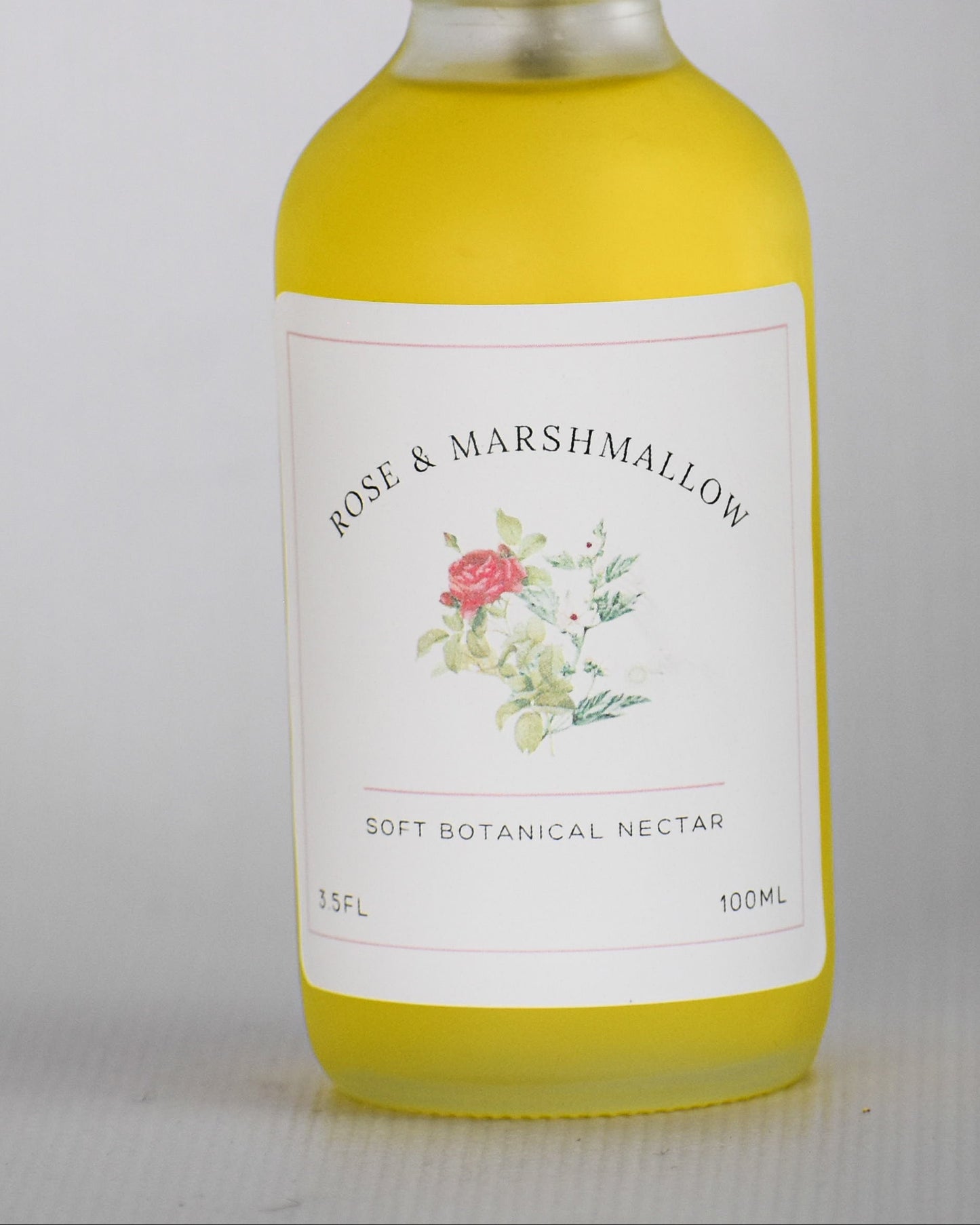 A bottle of Rose & Marshmallow Dry Botanical oil with a white spray top and a white label featuring a small floral illustration, filled with yellow liquid, set against a plain light gray background.