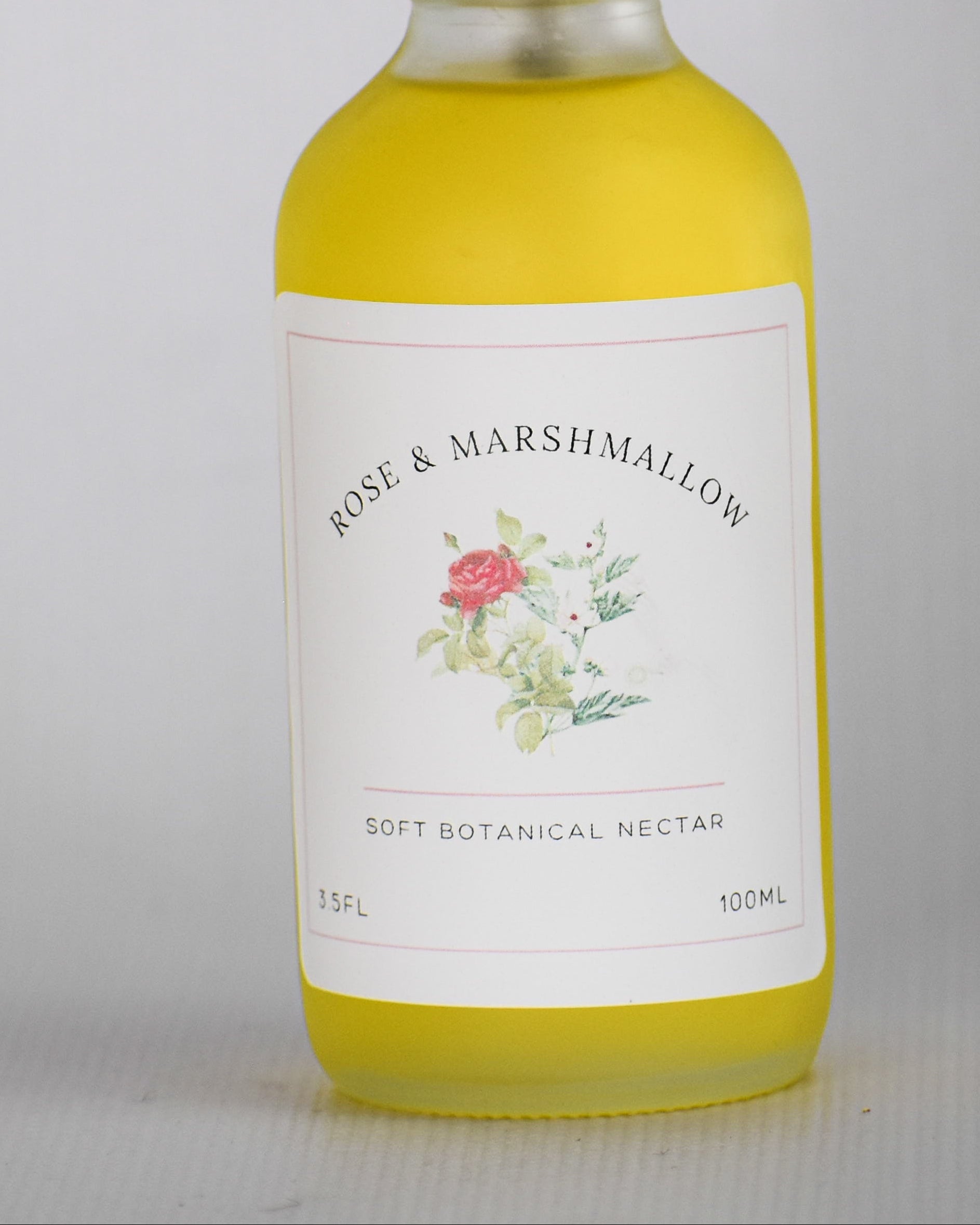 A bottle of Rose & Marshmallow Dry Botanical oil with a white spray top and a white label featuring a small floral illustration, filled with yellow liquid, set against a plain light gray background.