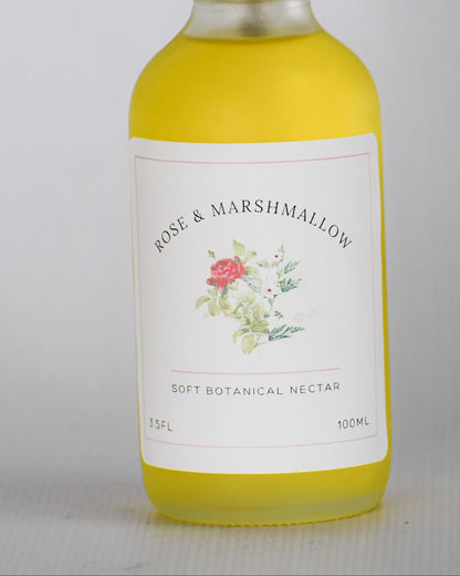 A bottle of Rose & Marshmallow Dry Botanical oil with a white spray top and a white label featuring a small floral illustration, filled with yellow liquid, set against a plain light gray background.