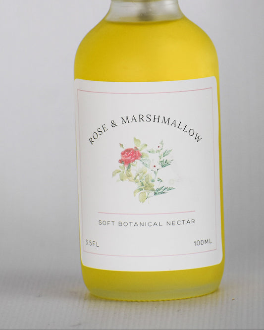 A bottle of Rose & Marshmallow Dry Botanical oil with a white spray top and a white label featuring a small floral illustration, filled with yellow liquid, set against a plain light gray background.